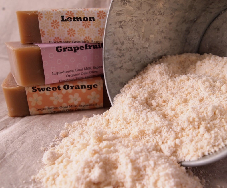 May include: Three bars of goat milk soap, stacked on top of each other. The bars are labeled "Lemon", "Grapefruit", and "Sweet Orange". The soap is spilling out of a metal container.
