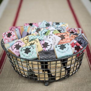 May include: A wire basket filled with small bars of goat milk soap. The soap bars are wrapped in colorful patterned paper with the words "gretta's goats" and "goat milk soap" printed on them.