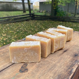 May include: A row of six rectangular bars of handmade soap, arranged on a wooden surface. The soap is a light brown color and has a slightly rough texture.