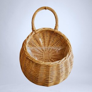 Könnte beinhalten: Ein runder, geflochtener Weidenkorb mit Henkel. Der Korb ist hellbraun und hat eine transparente Plastikauskleidung im Inneren. Der Henkel ist gewölbt und am oberen Teil des Korbes befestigt.