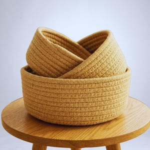 Könnte beinhalten: Drei runde, gewebte Körbe in verschiedenen Größen, die auf einem Holztisch gestapelt sind. Die Körbe sind in einem warmen Goldbraun gehalten und haben eine strukturierte, seilartige Optik. Der kleinste Korb befindet sich im mittleren Korb, der sich im größten befindet.