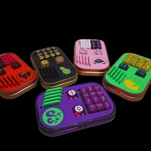 Könnte beinhalten: Fünf farbenfrohe Würfelboxen aus Metall, jede mit einem anderen Farbschema und Inhalt. Die Boxen enthalten Würfel, Spielsteine und anderes Spielzubehör. Die Boxen sind rot, schwarz, rosa, grün und lila.