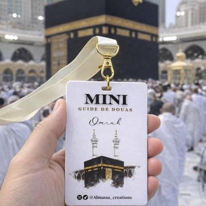 Puede incluir: Un cordón blanco con un cierre dorado que sostiene una tarjeta rectangular. La tarjeta presenta el texto "MINI GUIDE DE DOUAS Omrah" y una ilustración de la Kaaba. El fondo muestra una multitud de personas y edificios.