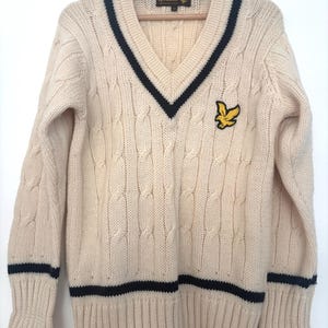 May include: A cream-colored cable knit V-neck sweater featuring dark navy blue trim along the neckline, cuffs, and hem. The garment displays a yellow embroidered eagle logo on the left chest and a Lyle & Scott brand label inside the collar.