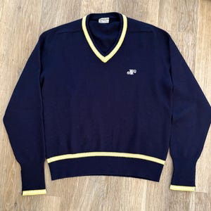May include: A navy blue long-sleeve sweater featuring a V-neckline with yellow trim. The sweater has a ribbed hem and cuffs with yellow stripes. A small white embroidered logo is positioned on the left chest area. A white label is visible at the neck.