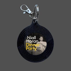 May include: A circular black keyring with a textured surface and a silver metal lobster clasp. The centre features a black and white photograph of Niall Horan with the text Niall Horan Dinner Party Live On Tour UK & Europe 2026 printed in white and yellow.