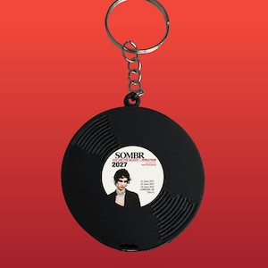 May include: A black circular keyring shaped like a vinyl record with a central white label. The label features the text SOMBR, YOU ARE THE REASON - WORLD TOUR, 2027, and a portrait of a person. It lists dates in June 2027 and LONDON, UK, The O2.