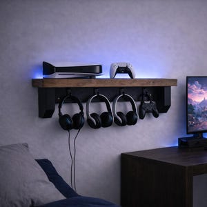 以下が含まれることがあります： ゲームセットアップ。木製の棚にコンソール、コントローラー、ヘッドフォンが置かれています。コンピューターモニター、キーボード、デスクランプが濃い木製のデスクにあります。部屋には青色のLED照明があります。