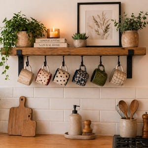 May include: A kitchen scene featuring a wooden shelf with hanging mugs of various designs. The shelf holds plants, a candle, and framed artwork. Below, a wooden cutting board and other kitchen accessories are visible, creating a cozy, rustic aesthetic.