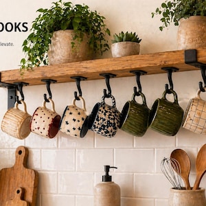 May include: A rustic wooden shelf with black iron hooks, displaying a collection of patterned mugs. The shelf is mounted on a white tiled wall, with potted plants above and below. Text on the image reads "UNDER SHELF IRON HOOKS Declutter. Organize. Elevate."