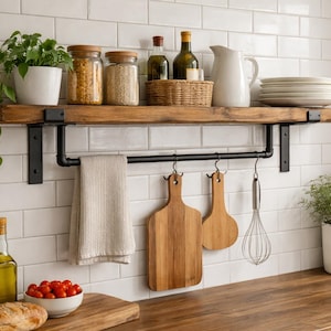 Puede incluir: Estante de cocina rústico con soportes de metal negro, con tarros de granos, una jarra blanca y platos. Debajo, un toallero con una toalla, tablas de cortar de madera y un batidor. Encimera de madera con tomates y pan.