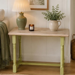 Op de afbeelding: Een kleine, rechthoekige houten bijzettafel met lichtgroene poten en een natuurlijk houten blad. Een brandende kaars, een lamp en een vaas met groen staan op de tafel. Een bank met een groen kussen en deken op de achtergrond.