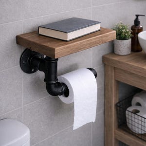 Puede incluir: Estante de baño rústico y portarrollos de papel higiénico. El estante es de madera oscura y está sostenido por tuberías de metal negro. Un libro gris descansa sobre el estante. Un rollo de papel higiénico blanco está en el soporte.