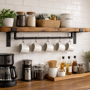 May include: A kitchen interior featuring a wooden shelf with jars, a coffee canister, and a woven basket. Below, a black rod holds white mugs. A coffee maker and grinder are on the counter, alongside bottles and a wooden tray.