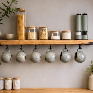 May include: A natural wood floating shelf with black metal brackets, featuring six hanging light grey speckled ceramic mugs. The shelf holds various glass storage jars with bamboo lids, two tall green grinders, and a beige ceramic vase with greenery.
