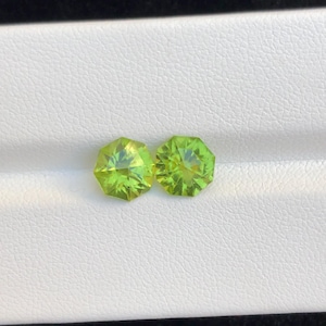 Puede incluir: Dos piedras preciosas de peridoto facetadas en forma de octágono. Las gemas son de un color verde claro y vibrante y se muestran sobre una superficie blanca. Las piedras preciosas tienen un corte brillante, que refleja la luz.