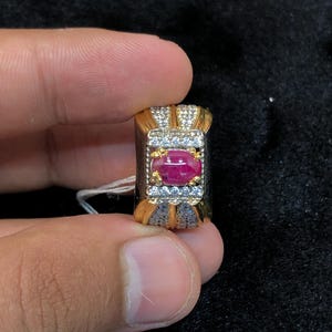 Peut inclure: Bague dorée avec un design rectangulaire orné d'un rubis ovale central et de petits diamants clairs. La monture de la bague présente un motif texturé. La bague est tenue par des doigts.