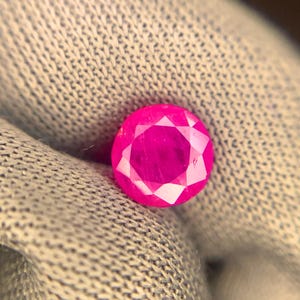 May include: A round, faceted, bright pink gemstone. The gem is set against a light grey, textured fabric. The facets of the gem reflect light, creating a sparkling effect. The gem is a vibrant, eye-catching colour.