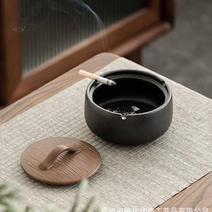 May include: A black ceramic ashtray with a lit cigarette, and a separate brown lid with a handle. The ashtray is on a woven mat on a wooden table. Smoke rises from the cigarette.