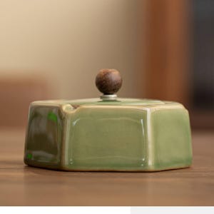 May include: A hexagonal, sage green ceramic ashtray with a small, round, wooden knob on the lid. The ashtray has a glossy finish and a small notch for resting cigarettes. The background is blurred, suggesting a focus on the ashtray.