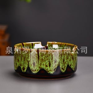 Könnte beinhalten: Ein Keramik-Aschenbecher mit grüner und brauner Glasur. Der Aschenbecher hat eine runde Form mit einem gewellten Rand und einem dunkelbraunen Boden. Der Text auf dem Aschenbecher ist auf Chinesisch.
