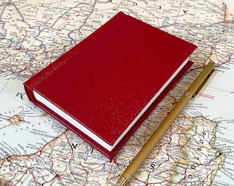 Puede incluir: Un cuaderno rojo de tapa dura descansa sobre un mapa antiguo, acompañado de un bolígrafo dorado. La cubierta del cuaderno tiene una apariencia texturizada. El mapa muestra características geográficas y nombres de lugares, sugiriendo un tema de viaje o planificación.