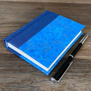 May include: A blue journal with a marbled design and a dark blue spine sits next to a black pen with silver accents. The journal's pages are white, and the cover is a vibrant blue.