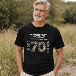 May include: Black t-shirt with white text that reads "Altersgerecht TOP in Schuss! Modelljahr 1956 - Legende in Serie." The number "70" is designed with gears and mechanical parts. The shirt is worn by a person.