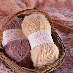 May include: A wicker basket holds two skeins of Juliet yarn, one light beige and one mauve. The yarn is labeled "Premium Quality Juliet". A small ball of yarn and knitting needles rest on a soft, pink, textured surface.