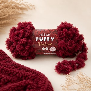 Könnte beinhalten: Ein Knäuel Alize Puffy FurLove Garn in einem tiefen Burgunderrot. Das Garn ist flauschig und strukturiert, mit dem Produktetikett sichtbar. Das Bild zeigt auch eine gestrickte Maschenprobe aus dem gleichen Garn, die sein weiches, plüschiges Aussehen zeigt. Das Garn ist für Projekte ohne Nadeln oder Haken konzipiert.