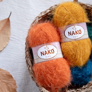 May include: Close-up of a woven basket filled with orange and yellow yarn skeins. Each skein has a label that reads "NAKO Paris". The yarn is fluffy and textured, suggesting a soft, warm material. A few dried leaves are visible.