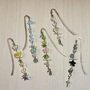 Puede incluir: Cinco marcadores de metal decorativos con colgantes. Cada marcador presenta un dise&ntilde;o diferente, incluyendo estrellas, flores, mariposas y otras formas caprichosas. Los colgantes son de varios colores, incluyendo amarillo, azul, verde y transparente.