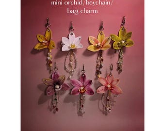 May include: Mini orchid keychains and bag charms in various colors, including yellow, white, pink, and purple. Each charm features a detailed flower design with beaded accents and a metal clasp. The text "mini orchid/keychain/bag charm" is at the top.