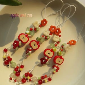 May include: Several beaded charms with red apple and flower designs. The charms have white cords and are adorned with red, green, and white beads. The text "apple and cat" is visible in the image.
