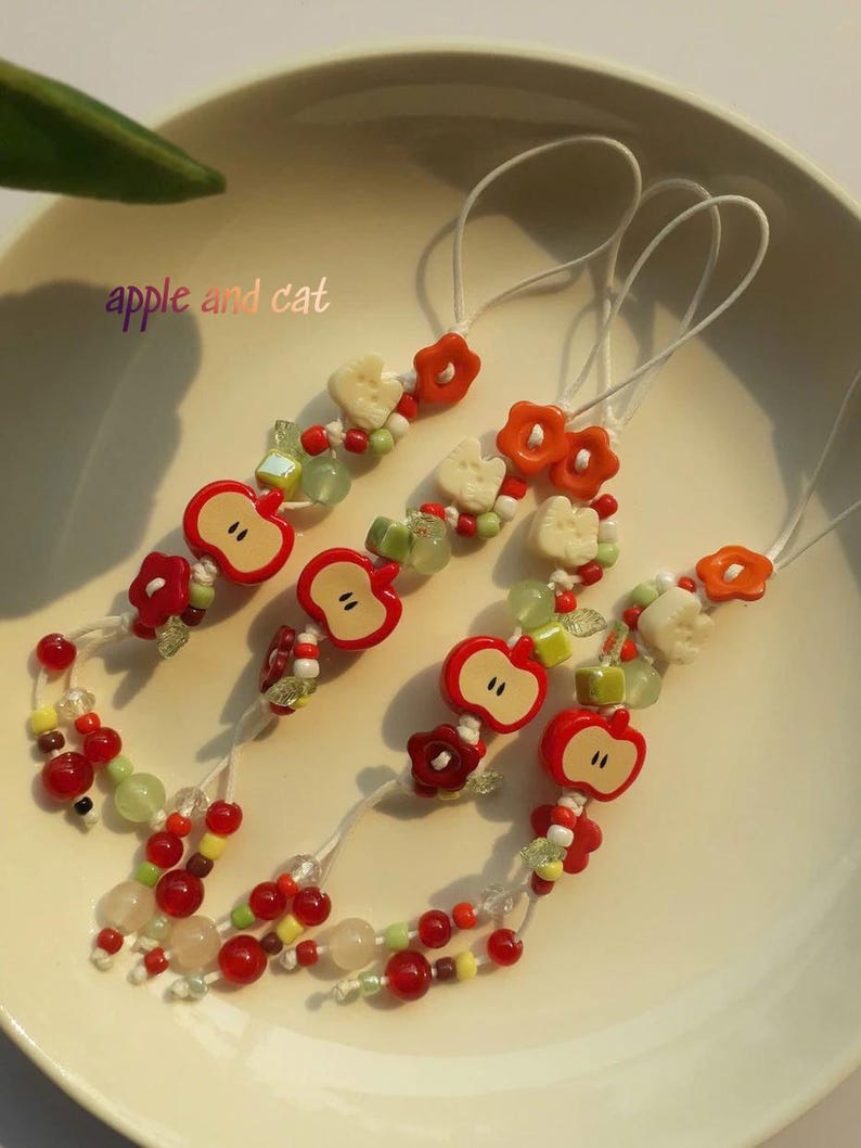 May include: Several beaded charms with red apple and flower designs. The charms have white cords and are adorned with red, green, and white beads. The text "apple and cat" is visible in the image.