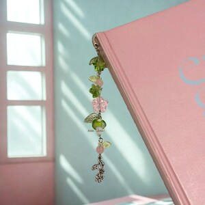 Puede incluir: Un marcap&aacute;ginas floral con cuentas y abalorios verdes, rosas y transparentes. El marcap&aacute;ginas est&aacute; unido a un libro rosa. Los abalorios incluyen hojas, flores y un peque&ntilde;o dise&ntilde;o floral plateado. El texto "#sea bread" es visible.