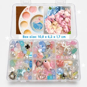 May include: A clear plastic storage box containing an assortment of colorful beads in various shapes, including stars, hearts, shells, and fish. The lid features images of a paint palette and pink and blue flowers, with text stating Box size: 10,8 x 6,2 x 1,7 cm.