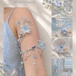 May include: A silver-toned arm cuff titled Moonlit Lily Arm Cuff, featuring a large lily flower, a crescent moon, and dangling charms including a star, small blue beads, and pearls. The design incorporates wire-wrapped elements and floral accents.