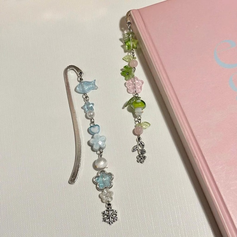 Puede incluir: Dos marcadores de libros con cuentas y detalles de metal plateado. Uno presenta peces azules, estrellas y dijes de copo de nieve. El otro tiene hojas verdes, flores rosas y un dije de hongo. Ambos est&aacute;n junto a un libro rosa.