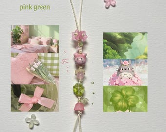 May include: A decorative hanging charm featuring a central string with pink and green beads, including a pink animal-shaped bead, a green clover-shaped bead, and small flower-shaped accents. The charm includes a loop at the top and two dangling ends.