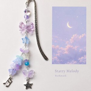May include: A silver metal bookmark with a curved, patterned spine, adorned with a dangling charm strand featuring purple and blue beads, translucent butterfly shapes, star-shaped beads, flower-shaped beads, a music note charm, and a star charm.