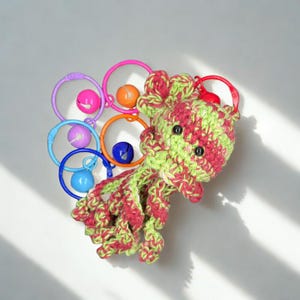 May include: A handmade crochet octopus toy in shades of green and red, with black button eyes. Attached are colorful plastic rings with bells in pink, orange, blue, purple, and red. The toy is designed for babies and young children.