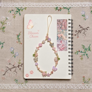 May include: A phone charm featuring a beige cord strung with various beads in shades of pink, purple, and white. The beads include shapes of butterflies, flowers, stars, and hearts, along with round pearls. The charm is displayed on a white notebook.