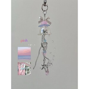 May include: A pink and silver keychain featuring a clear bell-shaped charm, adorned with a bow, beads, and a starfish pendant. The word "pink" is written in pink. Includes small images of a pink sunset and a pink building.