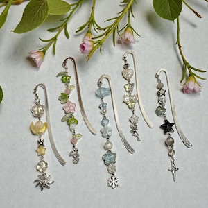 May include: A collection of five decorative metal bookmarks with dangling charms. Each bookmark features a different design, including flowers, stars, butterflies, and beads in various colors like pink, green, blue, and yellow. The bookmarks are displayed against a soft, light background.
