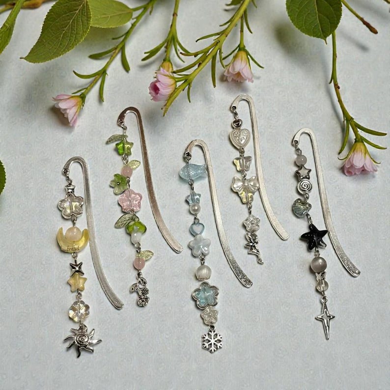 Puede incluir: Colecci&oacute;n de cinco marcap&aacute;ginas decorativos de metal con colgantes. Cada marcap&aacute;ginas presenta un dise&ntilde;o diferente, con flores, estrellas, mariposas y cuentas en varios colores como rosa, verde, azul y amarillo. Los marcap&aacute;ginas se muestran sobre un fondo suave y claro.