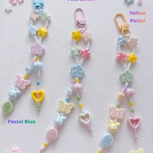 May include: Three beaded keychains featuring pastel-colored charms including flowers, butterflies, hearts, and animal faces. Each keychain has a lobster clasp and small white seed beads. Text labels read Pastel Blue, Pink Sweet, and Yellow Pastel.