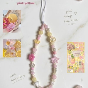 May include: A beaded phone charm in pink, yellow, and white. The charm features various beads, including pearls, stars, flowers, and a seashell. The words "pink yellow", "good things take time", and "little happiness" are also visible.