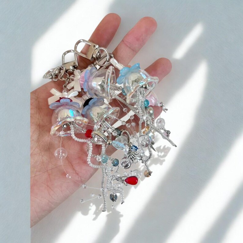 May include: A collection of clear, bell-shaped charms with iridescent accents, adorned with small bows, beads, and silver-toned metal details. The charms are attached to a silver keychain, creating a whimsical accessory.