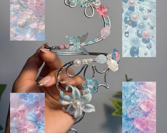 May include: A silver-toned wire ear cuff titled Mermaid Whisper, featuring iridescent flower-shaped charms, small pink and white beads, and a translucent butterfly accent. The design includes swirling wire elements and delicate floral components.